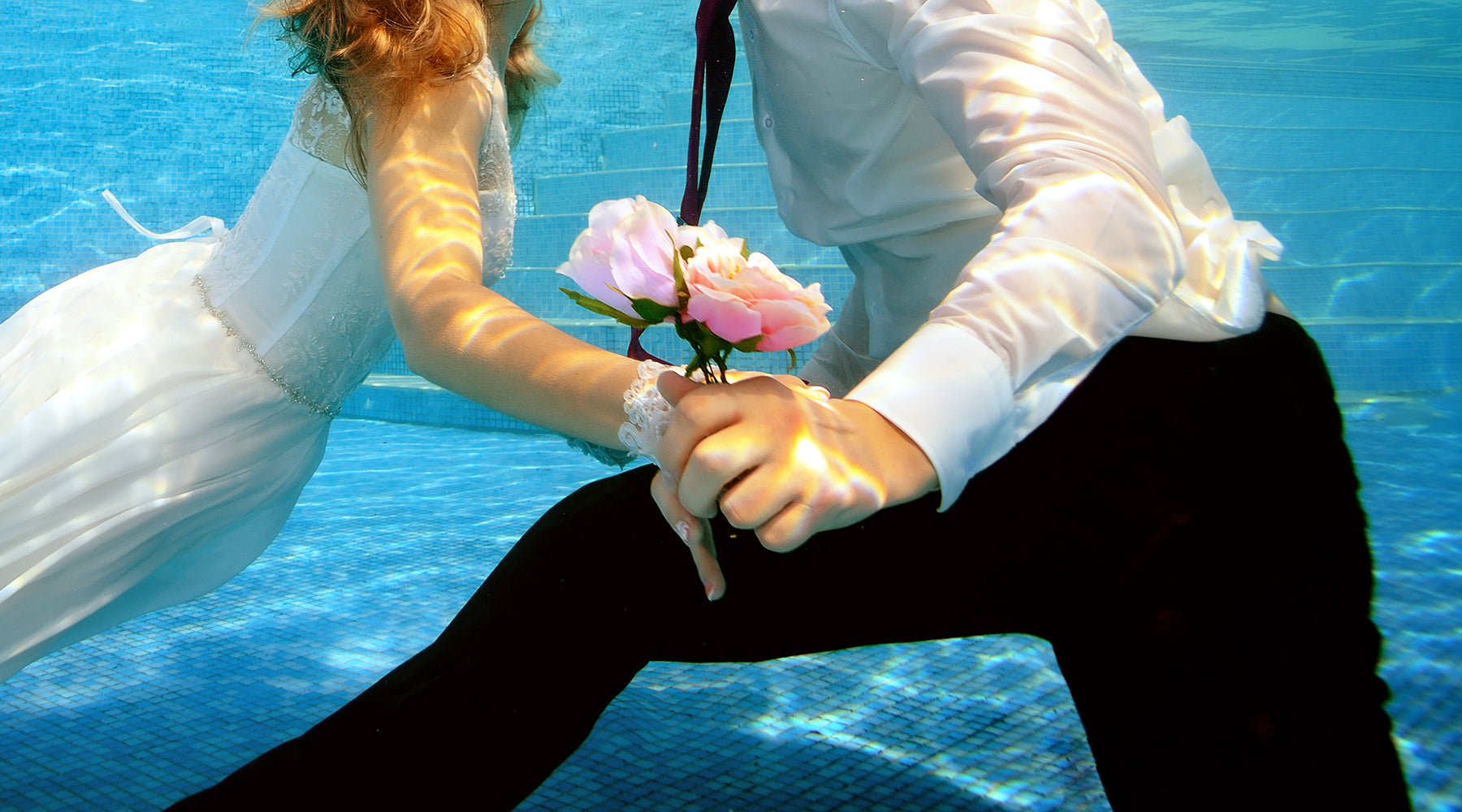 A bride in a white dress and groom in a white shirt with black pants hold a pink bouquet as they kiss under the water of a pool.